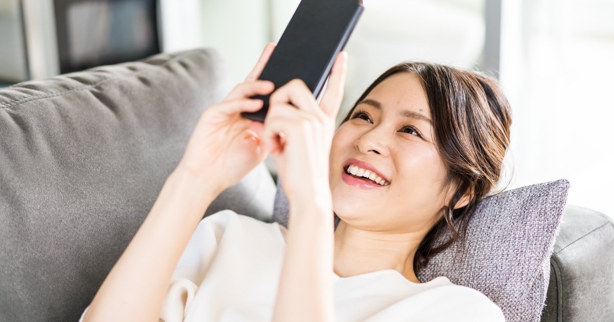 ソファに横になり、スマホを両手で持って笑う女性
