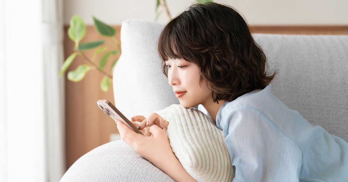 ソファのクッションにもたれ、スマホの画面を読む女性