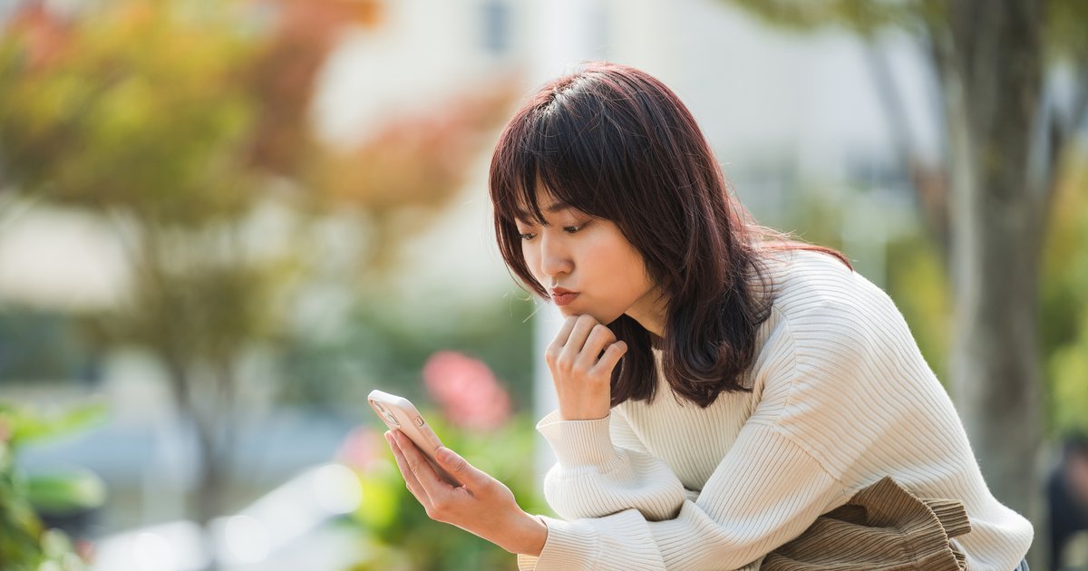 屋外で前かがみになり、顎に手を当ててスマホを見る女性