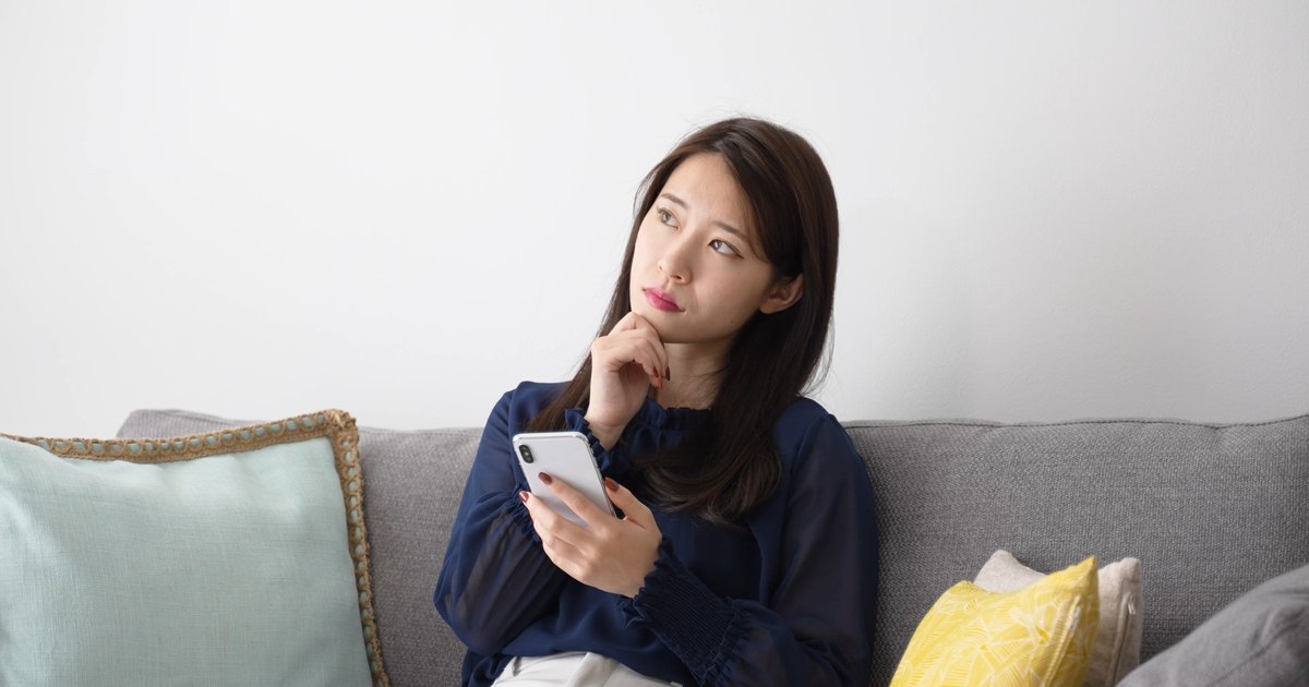 ソファでスマホを持ち、口元に指を当てて考える女性