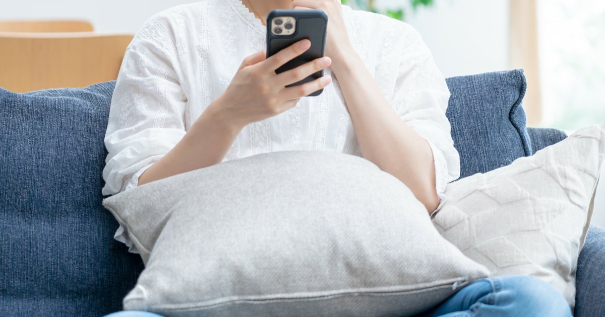 ソファに座り、クッションを膝に置いてスマホを手に持つ人の上半身
