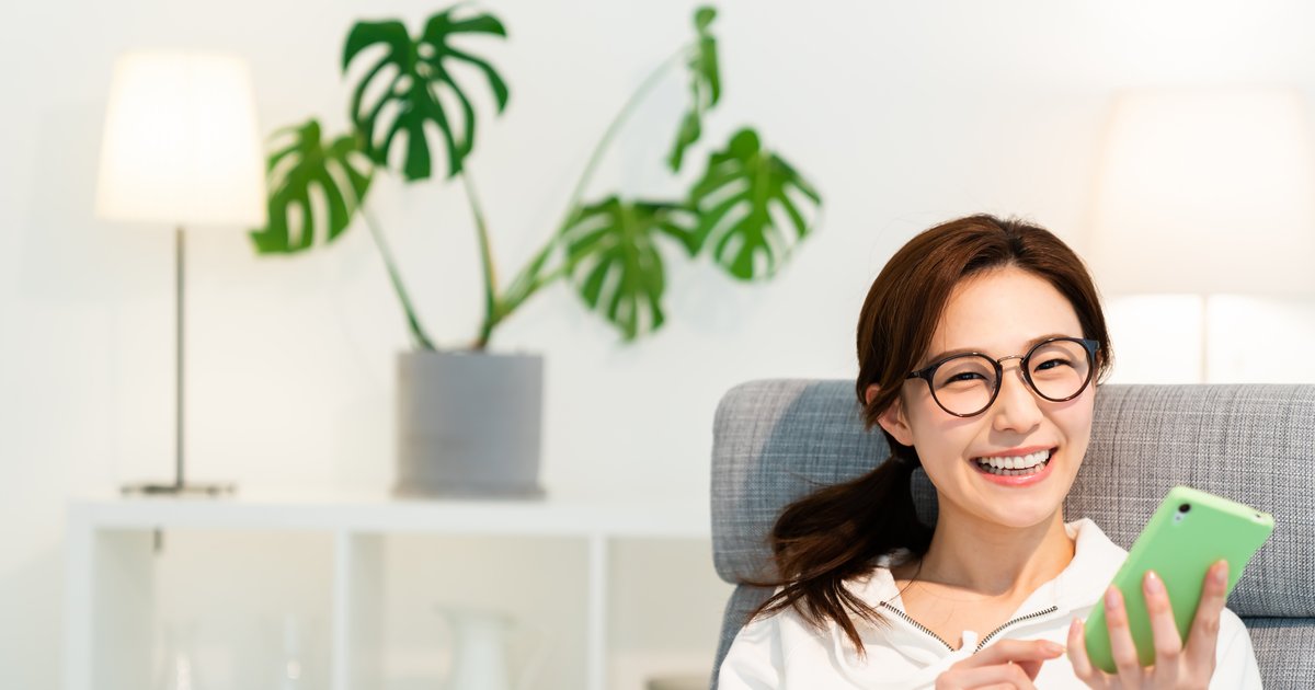 観葉植物のある明るい部屋で、スマホを手に笑う眼鏡の女性