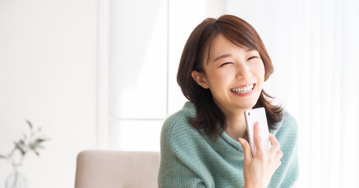 明るい部屋でスマホを頬の近くに持ち、目を閉じて笑う女性