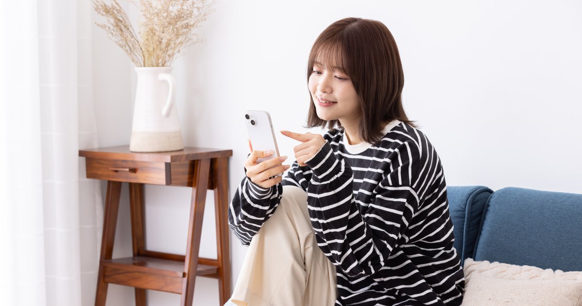 ソファに座り、スマホの画面を指さしながらアプリを操作する女性