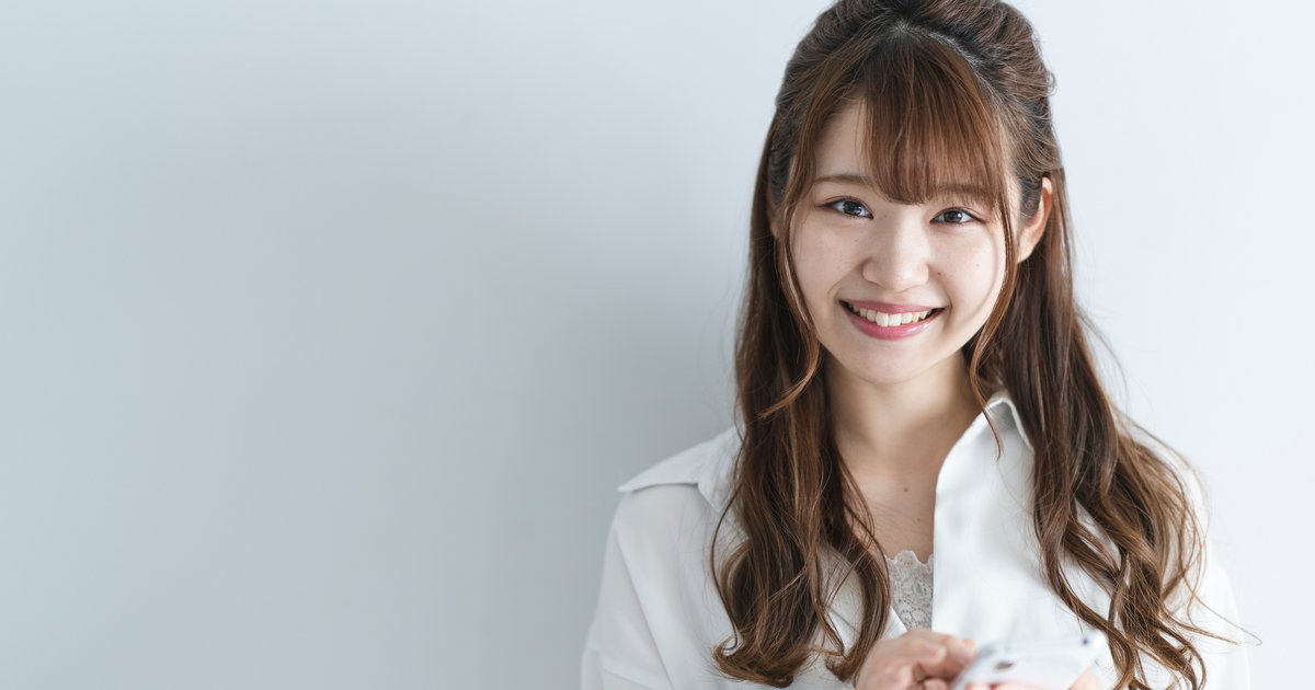 白い壁を背にスマホを両手で持ち笑顔の女性