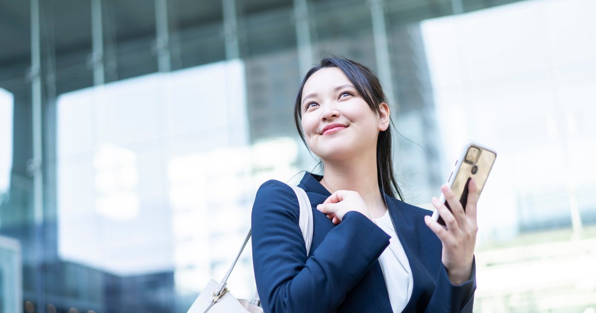 オフィス街でスマホを手に持ち、笑顔で見上げるスーツ姿の女性