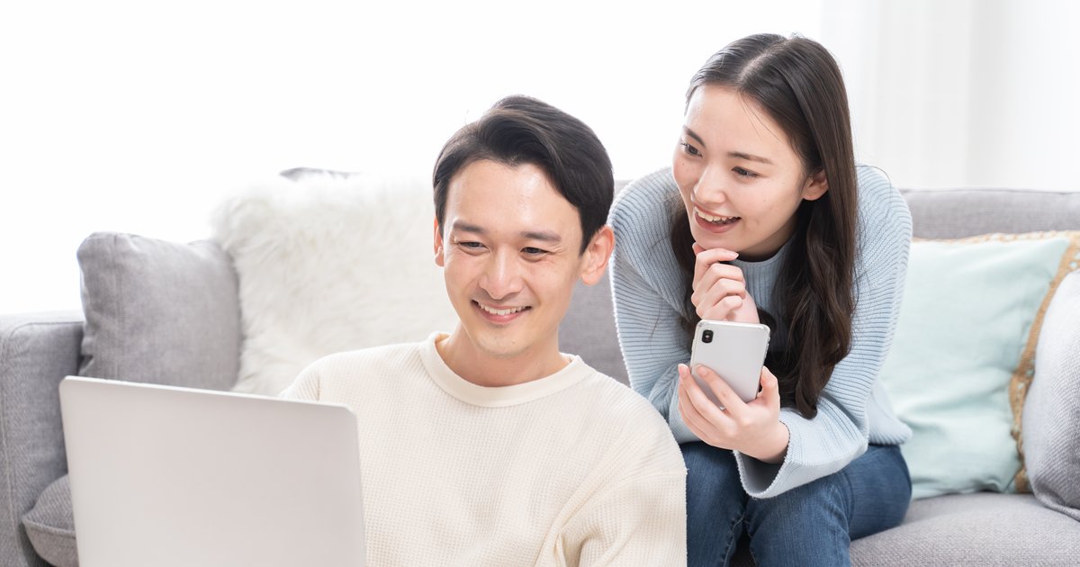 ソファのある部屋で、ノートパソコンの前で笑う男性と、スマホを手に笑う女性