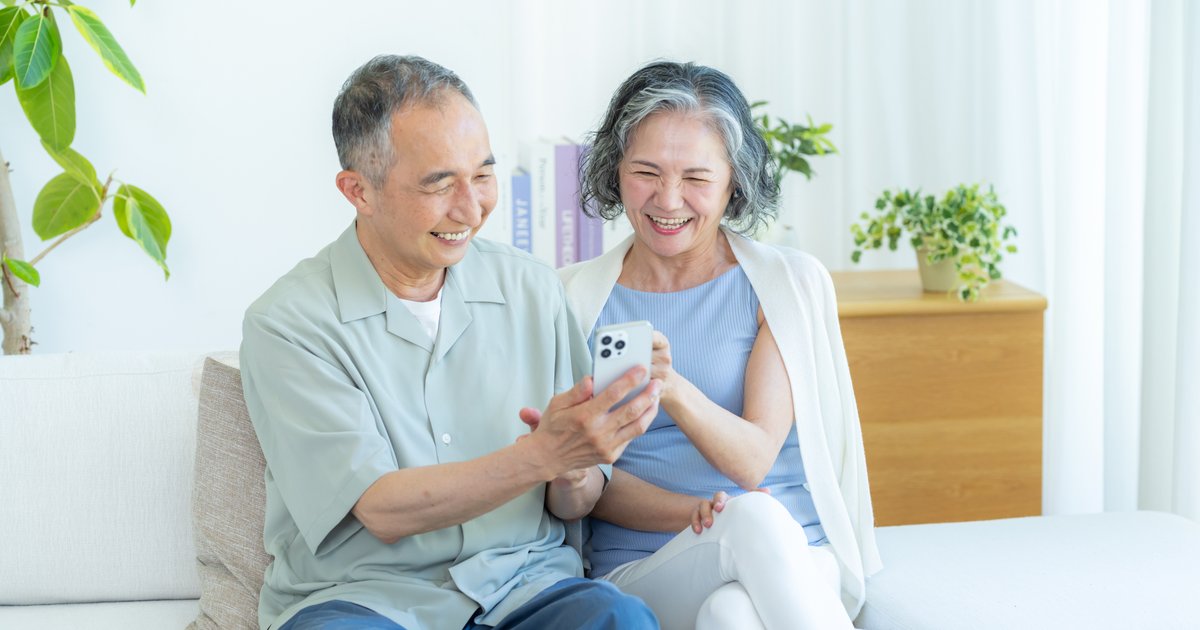 ソファに並んで座り、1台のスマホを手に笑い合う年配の男女