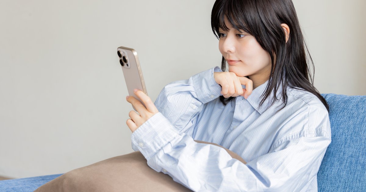 ソファで頬に手を当て、スマホを見ながら考える女性