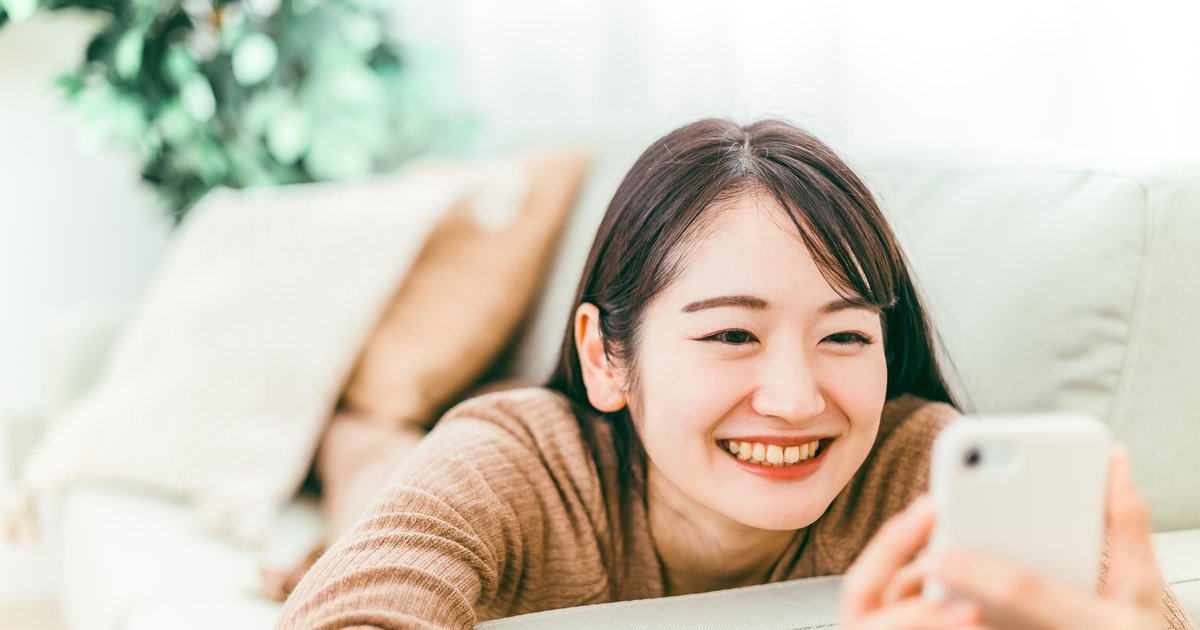 ソファに横になり、スマホの画面を見て笑顔になる女性