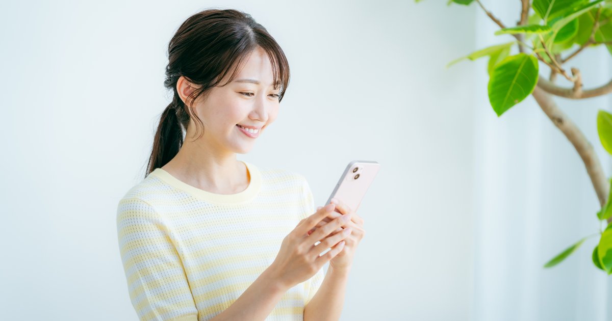 明るい部屋で観葉植物のそばに立ち、スマホを見て微笑む女性