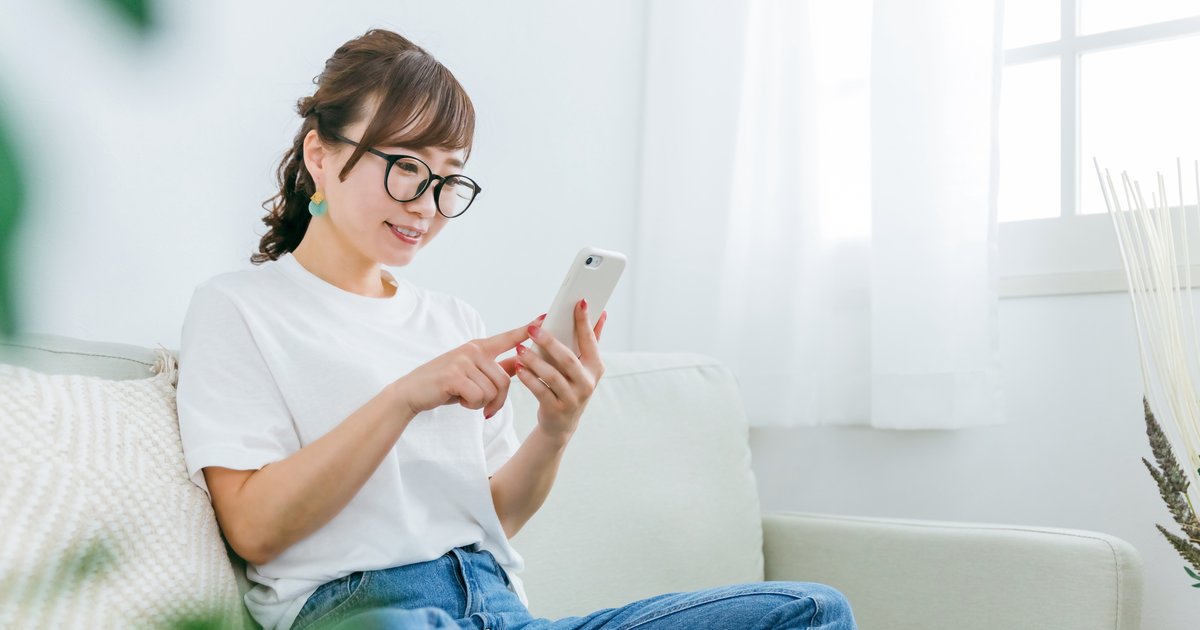 明るい部屋のソファに座り、スマホの画面を指でタップする女性